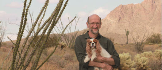 Bill Langer standing in the Arizona desert holding a small dog, with ocotillo and a rocky ridge behind him.